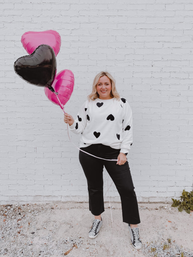 Valentine's Inspired Looks + Gifts + Decor Ideas - Woman holding balloons in white sweater with black hearts, black pants and sneakers in front of brick wall